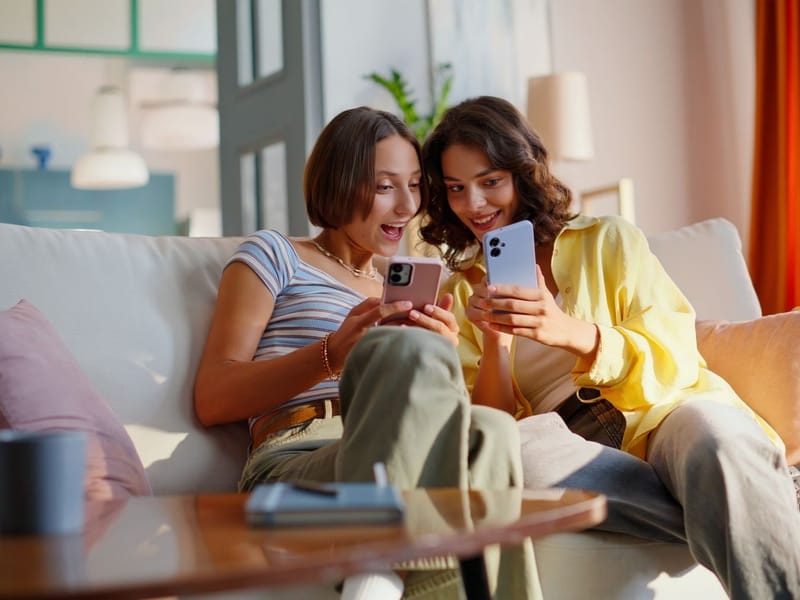 Zwei junge Frauen sitzen auf dem Sofa und schauen lachend gemeinsam auf ihre Smartphones